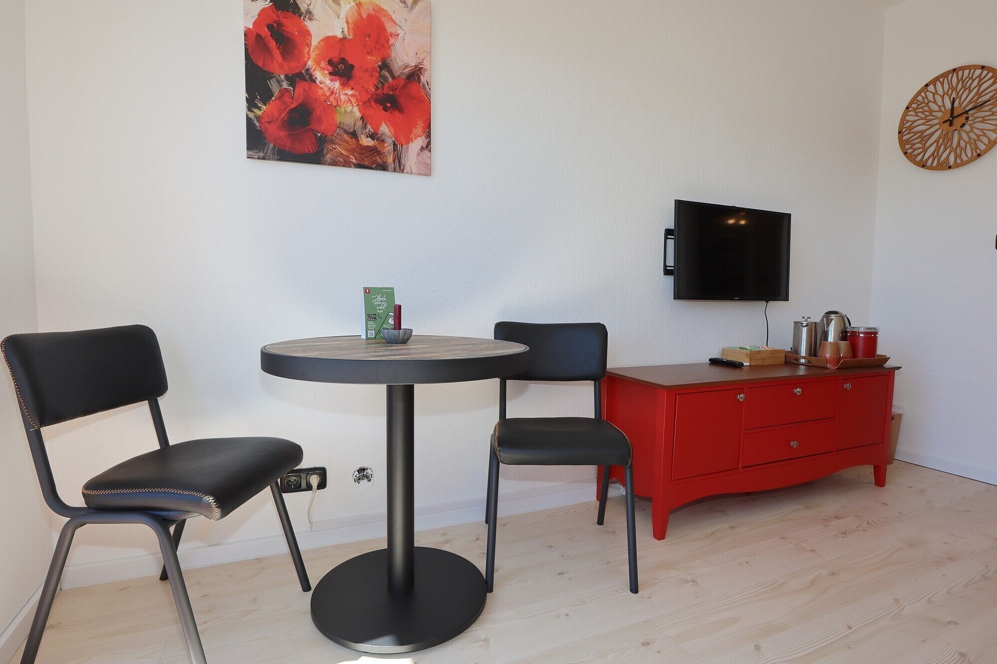 Une pièce comprenant une table ronde, deux chaises, un buffet rouge, un téléviseur à écran plat et un tableau accroché au mur.