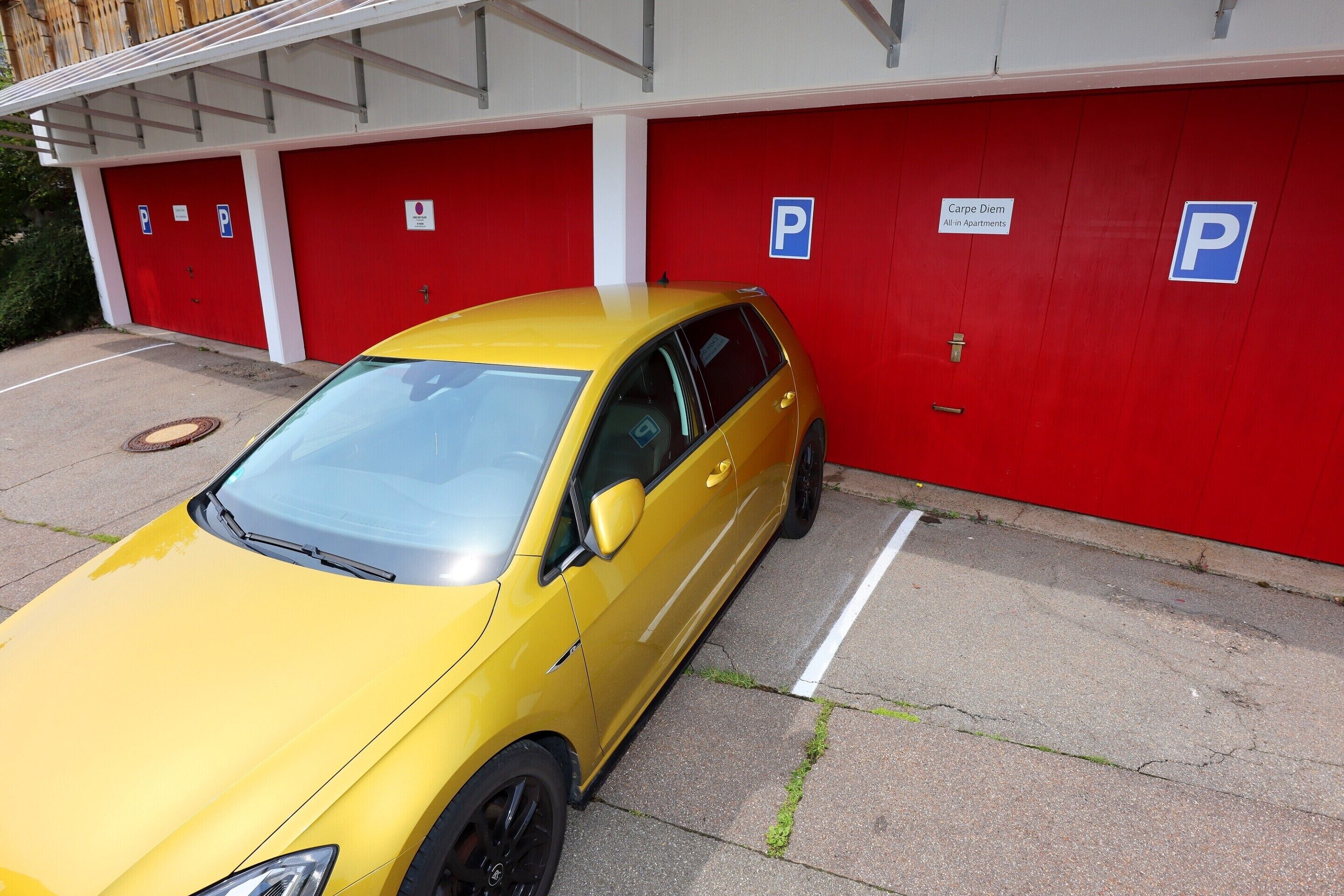Un coche amarillo estacionado frente a un edificio rojo y blanco con varios espacios de estacionamiento y un cartel que dice 'Carpe Diem All in Apartments'.