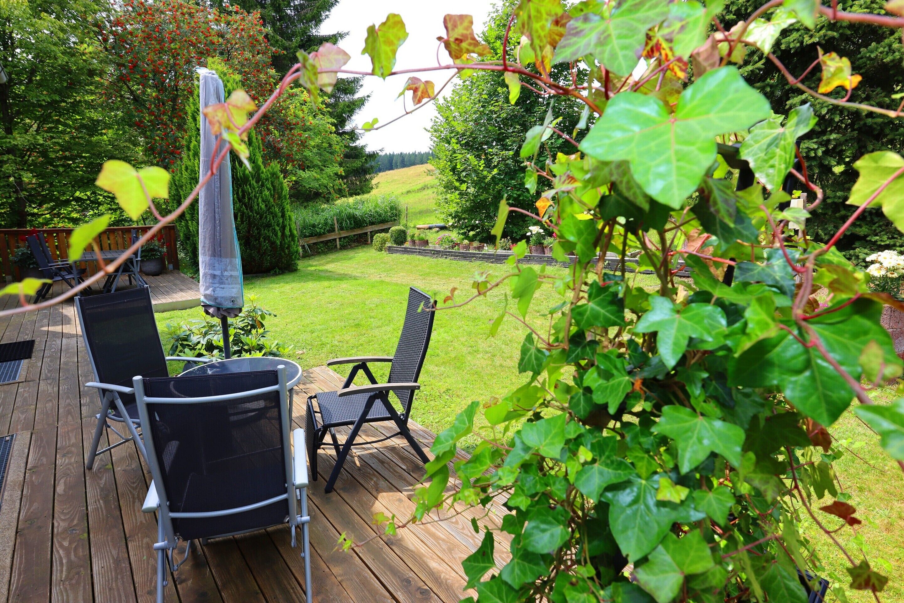 A wooden deck with two black chairs and a table, surrounded by lush greenery and a well-maintained lawn.