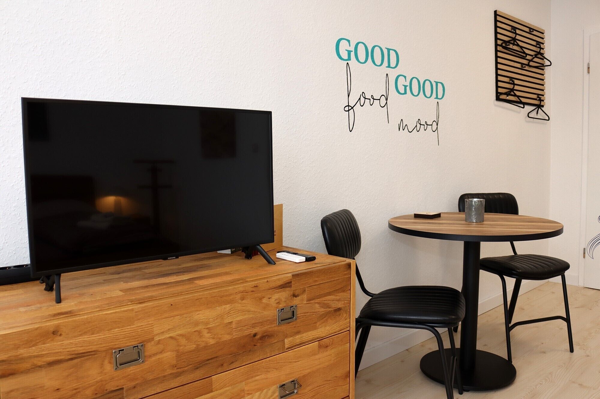 A room with a TV, wooden dresser, two black chairs, a small round table with a cup, and a wall with 'GOOD food GOOD mood' decor.