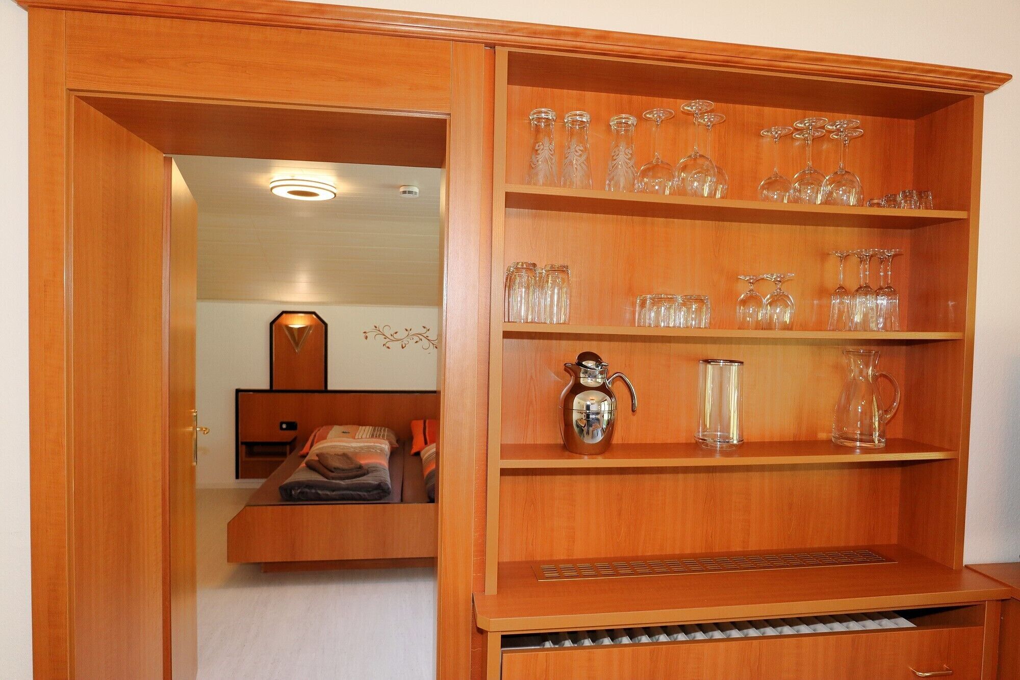 A wooden cabinet with shelves holding various glassware and a silver pitcher.