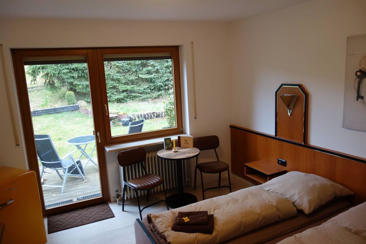 A bedroom with a bed, a small table, two chairs, a radiator, and a window with a view of the outdoors.