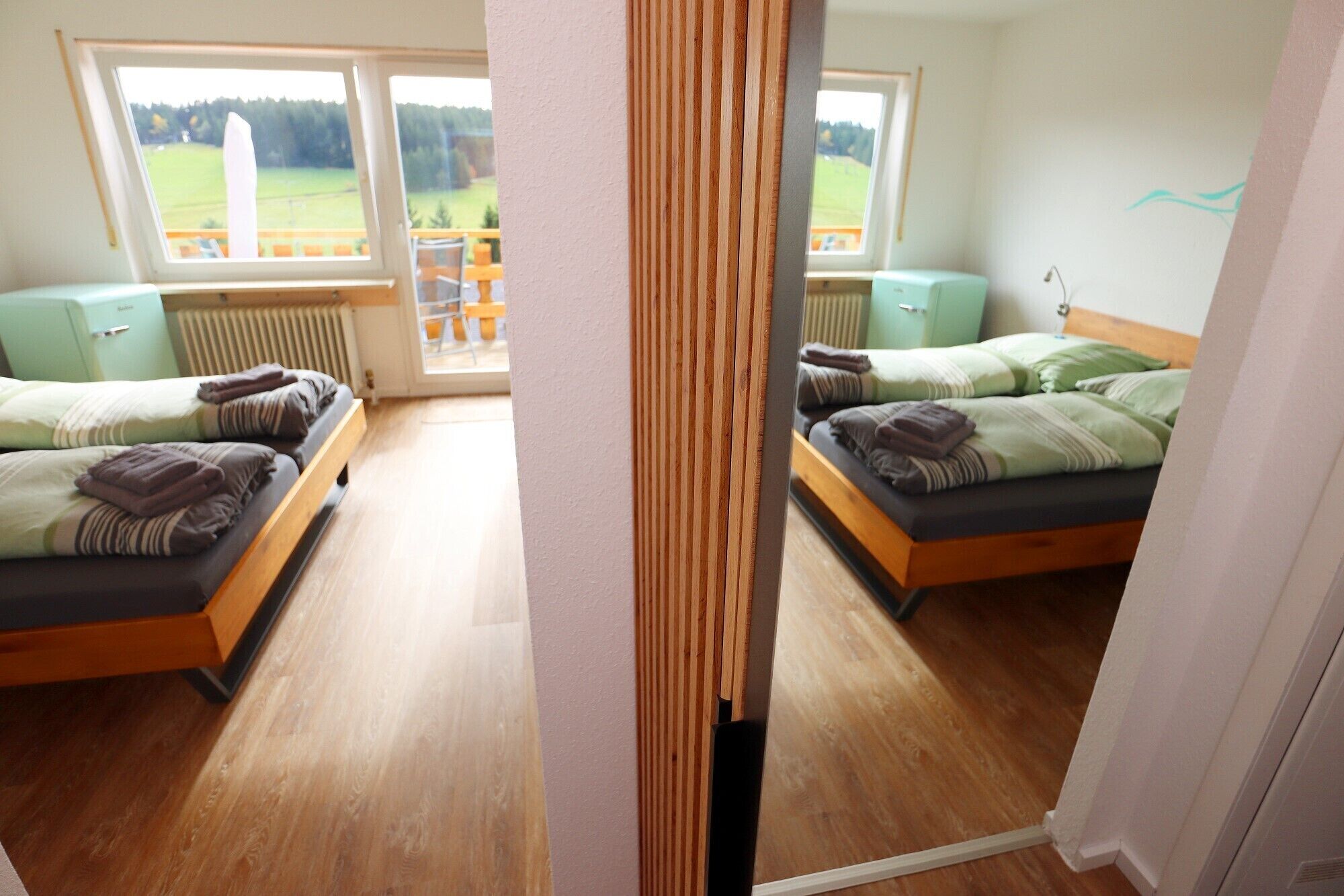Une chambre d’hôtel avec deux lits, un plancher en bois, une vue sur un paysage verdoyant par une grande fenêtre, et une petite table avec des chaises à l’extérieur.