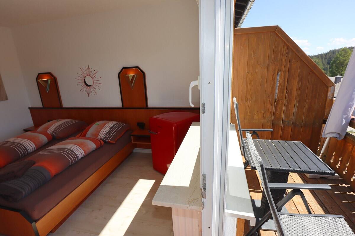 A bedroom with a bed, a red bench, a wooden wall, and a view of the outdoors.