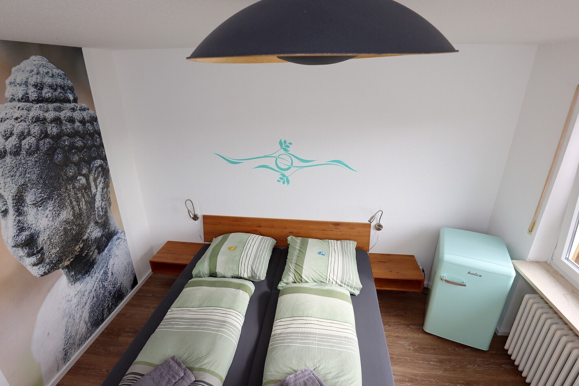Une chambre avec un lit, une lampe fixée au mur, un radiateur et un petit réfrigérateur.