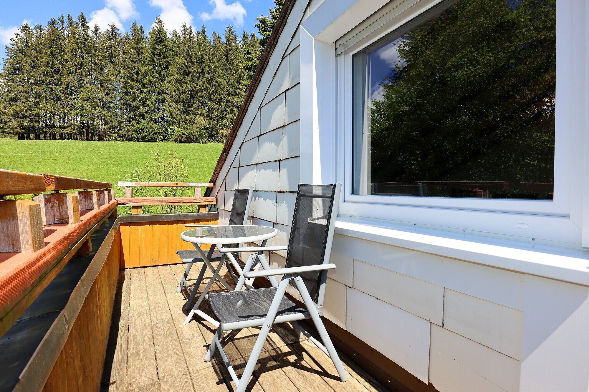 En træterrasse med et bord og stole, et vindue med udsigt over en skov, og en hvid facade.