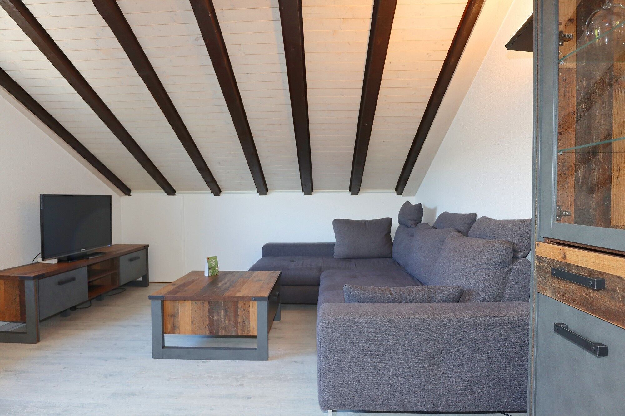 Uma sala de estar moderna com teto de madeira, um sofá cinzento, uma mesa de centro e uma televisão.