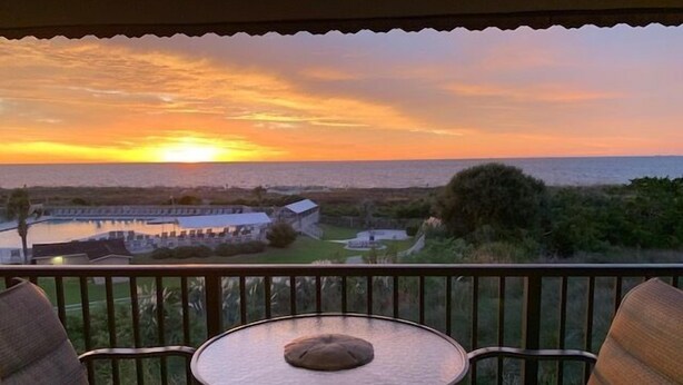 Un balcon avec une table ronde et deux chaises, donnant sur une étendue d’eau et une côte lointaine, au moment du coucher du soleil.