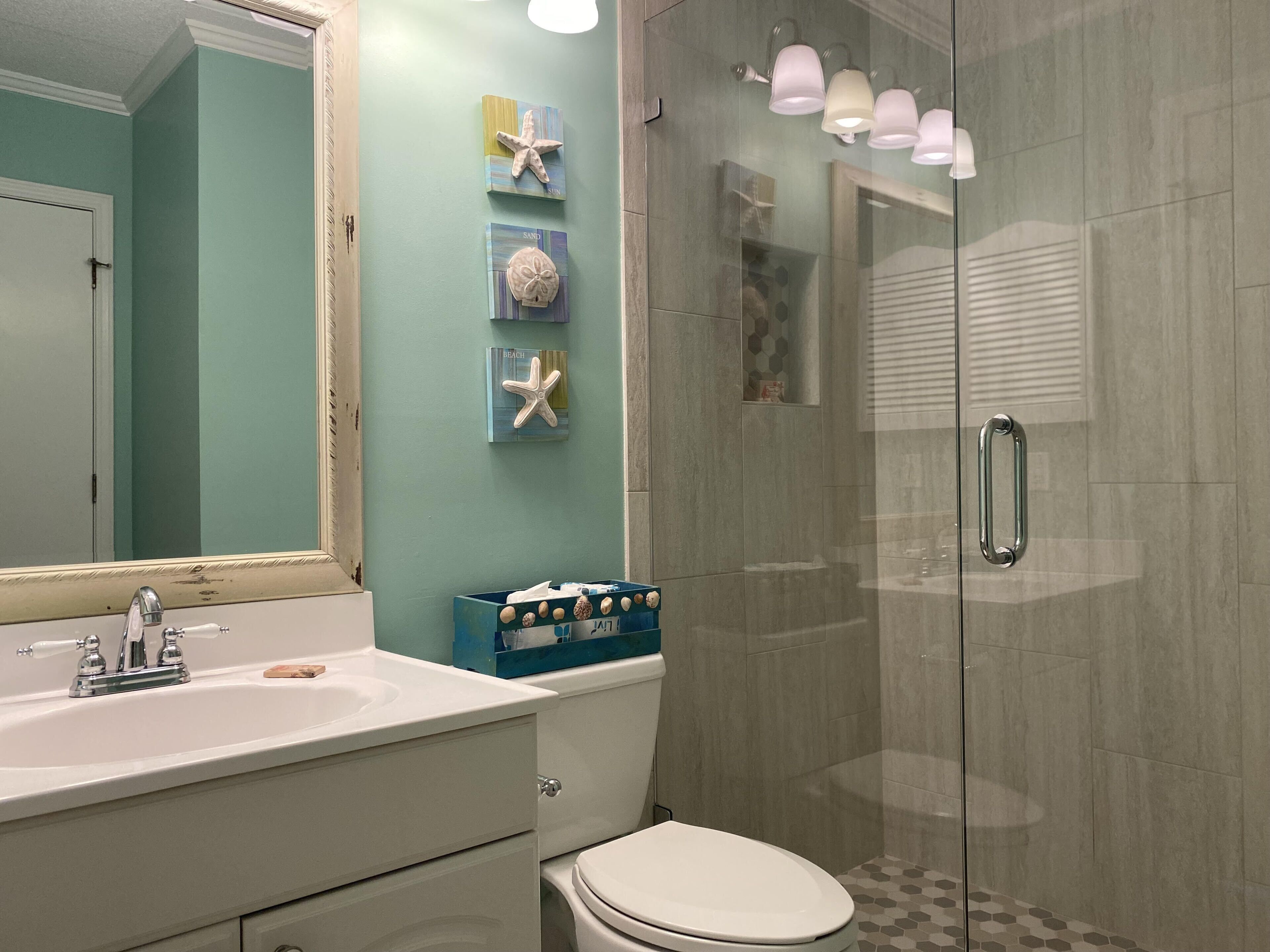 Un baño con un lavamanos blanco, un inodoro y una ducha con mampara de vidrio. Se observan decoraciones de estrellas de mar en la pared y un estuche de almacenamiento azul.