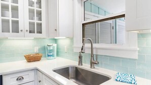 Eine Küche mit weißen Schränken, einer Edelstahlspüle, einem Fenster mit Jalousien und einer hellblauen Spritzschutzleiste.