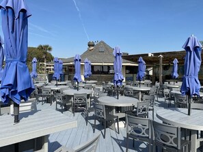 Ein Außenbereich mit runden Tischen und Stühlen, beschattet durch blaue Sonnenschirme, und ein Gebäude mit geneigtem Dach im Hintergrund.