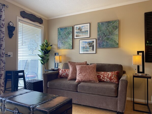 A living room with a brown sofa, two cushions, a small table, a lamp, and a wall with three framed pictures. - Resort View - Ocean Villas C206-Professionally Managed by HHIB&T Vacation Rentals (Hilton Head Island)