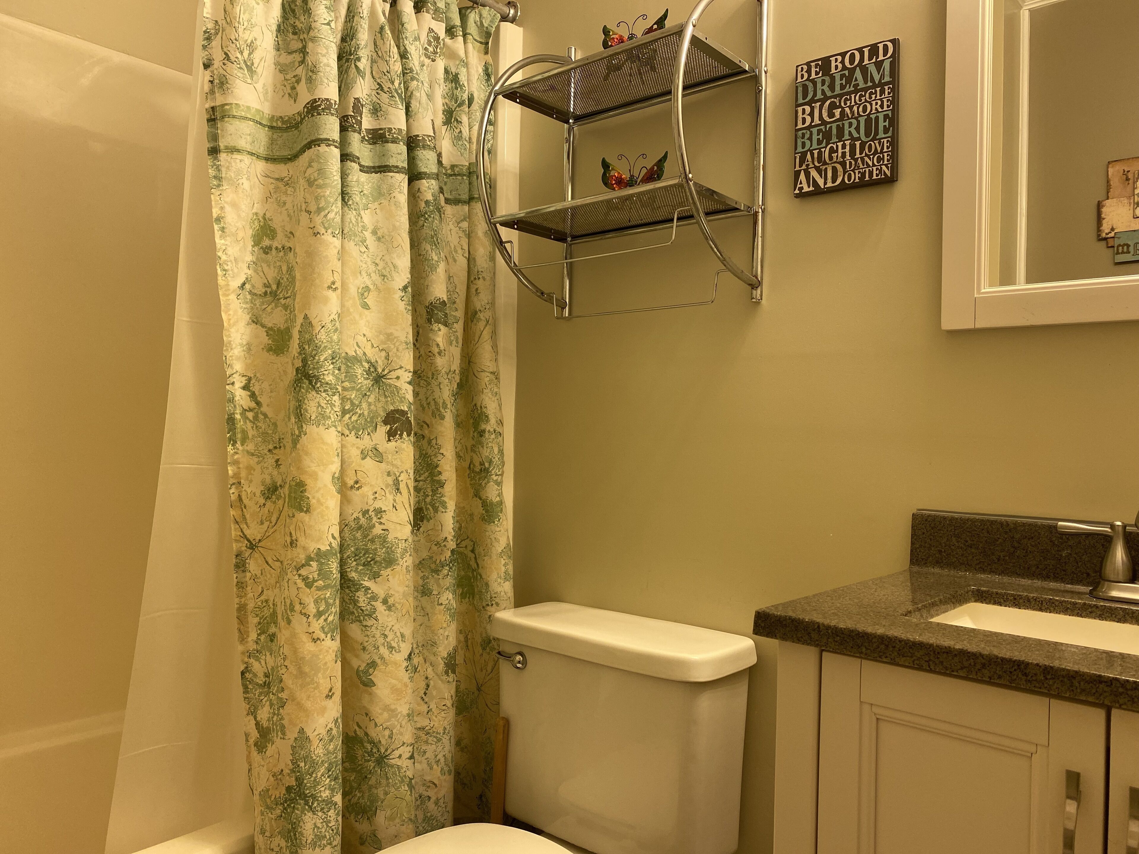 A bathroom with a toilet, a shower curtain, a shelf with decorative items, and a motivational quote on the wall.