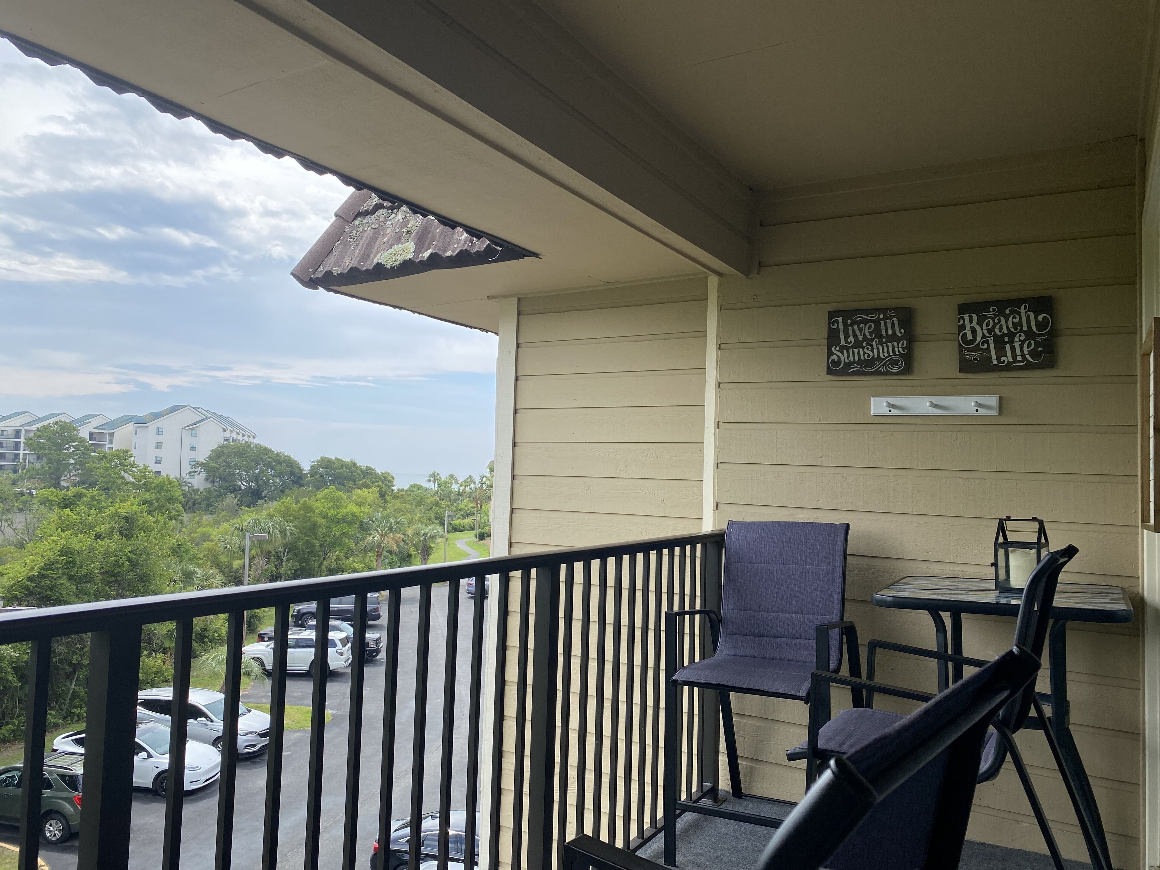 A balcony with a view of a parking lot and buildings, a chair, a small table, and two signs on the wall.