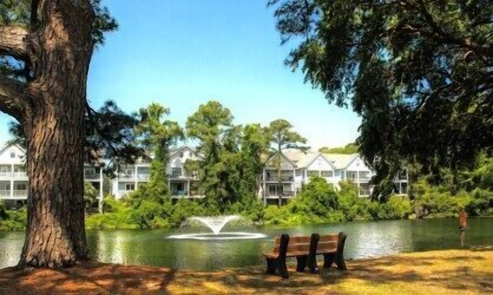 A lakeside view with a bench, trees, and a building complex in the background.