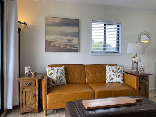 Een woonkamer met een bruine leren bank, een houten bijzettafeltje, een lamp en een lijst met een foto van een strandgezicht.