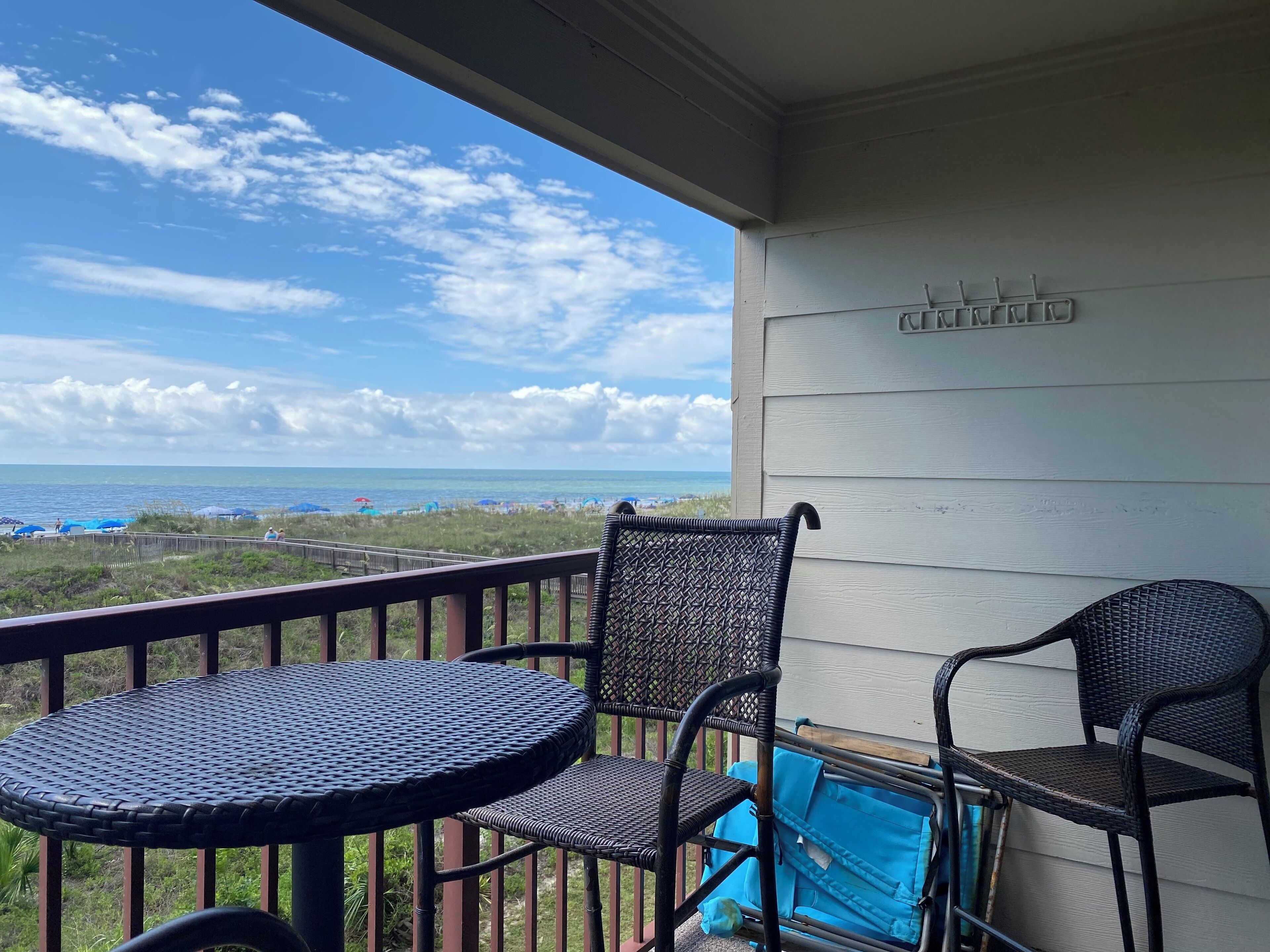 A balcony with a table and two chairs, overlooking a beach with umbrellas and the ocean.