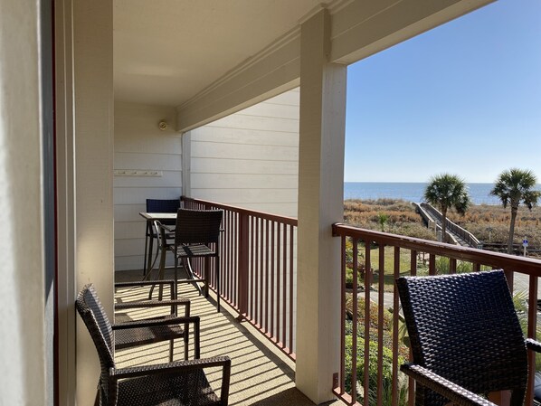 A balcony with a table and chairs, overlooking a beach with palm trees and a clear sky.