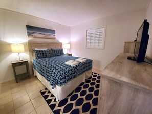 A bedroom with a bed, two bedside tables, a lamp, a painting, a window with shutters, and a desk with a computer. - Ocean View - Admiral’s Row 319-Professionally Managed by HHIB&T Vacation Rentals (Hilton Head Island)