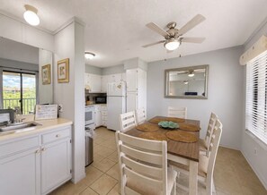 Una cocina compacta con armarios blancos, una mesa de comedor con sillas, un ventilador de techo y una ventana con persianas.
