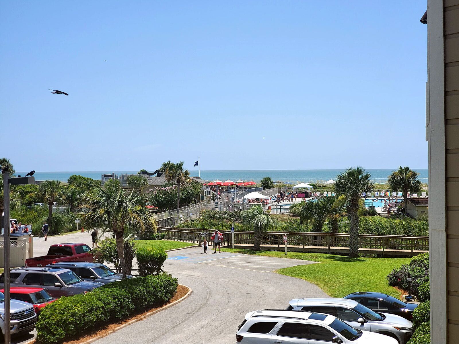 Vista de un estacionamiento con coches, un área de piscina y una playa al fondo.
