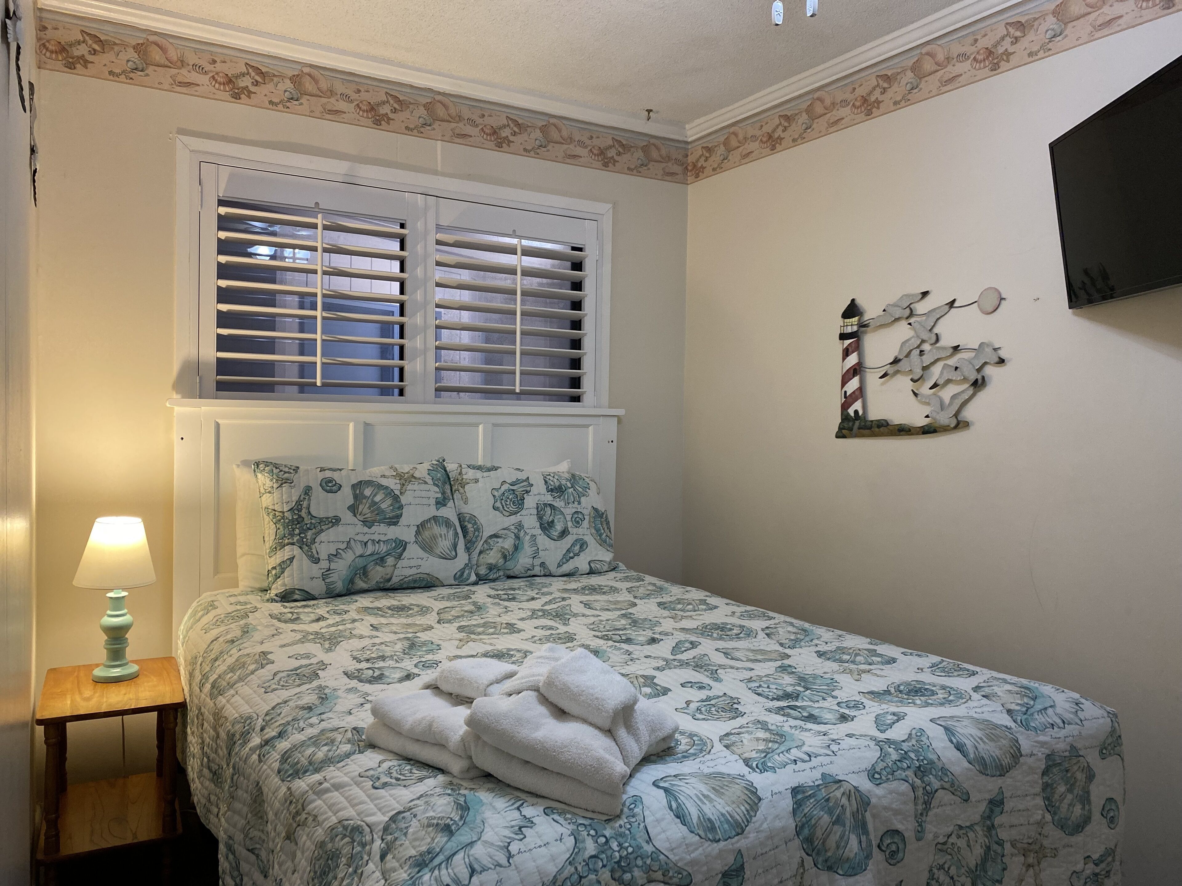 Une chambre à coucher comprenant un lit, une table de chevet, une lampe, une fenêtre avec des volets et un élément de décoration murale.