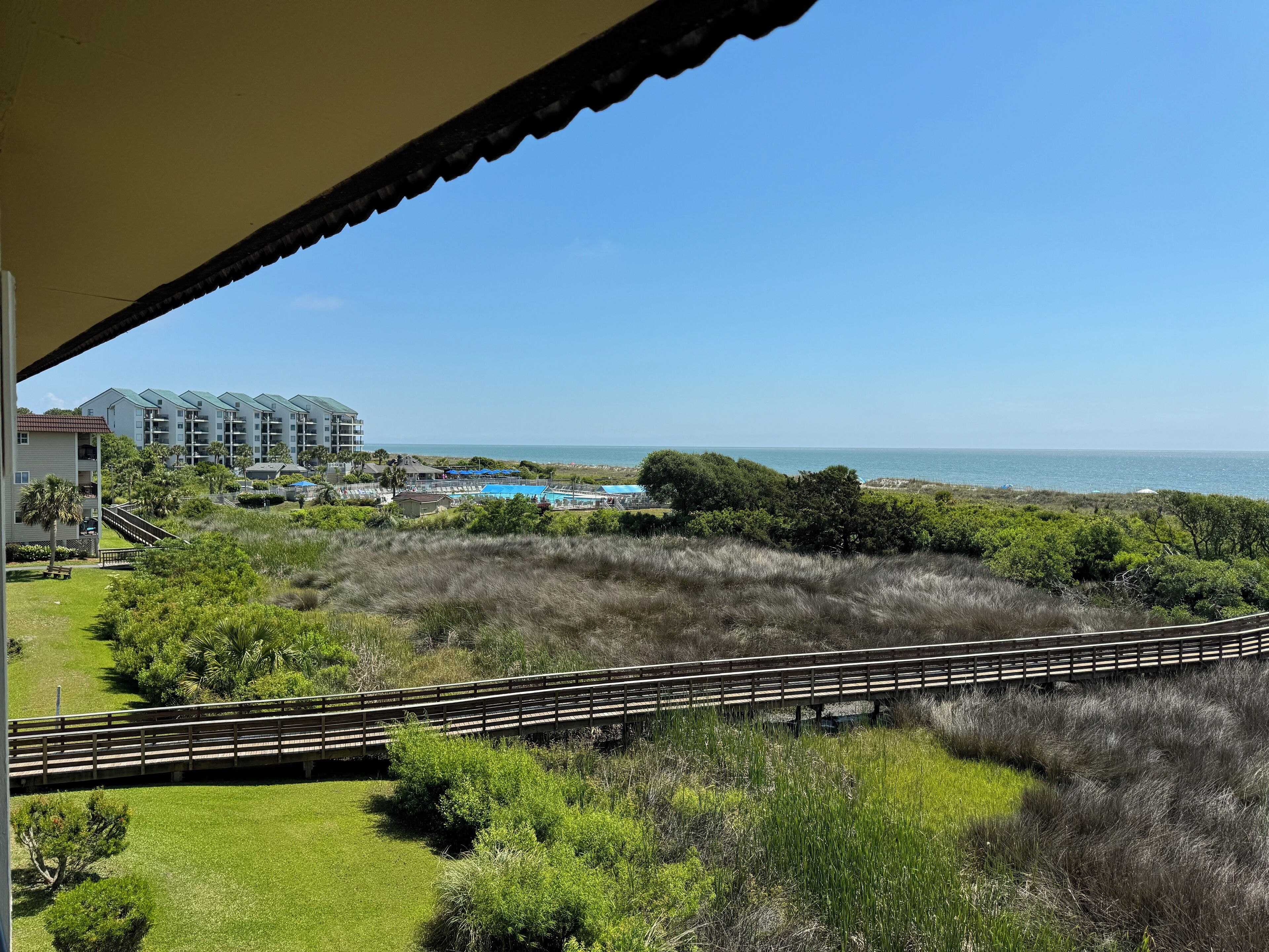 A view of a coastal hotel with a swimming pool, lush greenery, and a clear blue sky.