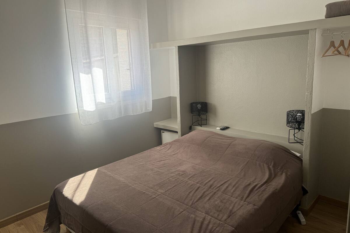 Un dormitorio con cama, mesita de noche, lámpara y ventana con cortina translúcida.