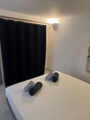 Ett rum med en säng, mörka gardiner, en väggmonterad lampa och hoprullade handdukar på sängen.
