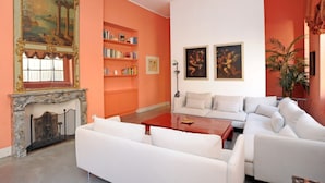Een woonkamer met een open haard, witte banken, een rode salontafel en boekenplanken tegen een oranje wand.