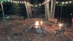 A campfire with a bench and chairs around it, surrounded by trees and string lights.
