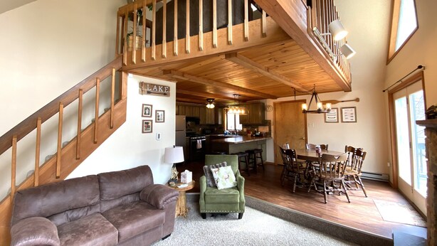 A spacious living area with a brown leather sofa, a green armchair, a wooden staircase, a dining area with wooden chairs, and a kitchen in the background.