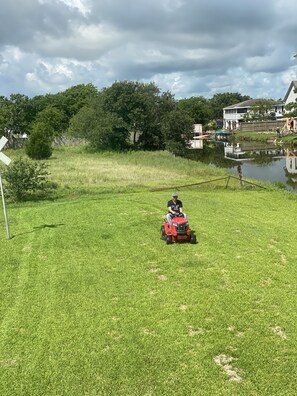 A person riding a red lawn mower on a grassy field with a pond and trees in the background.