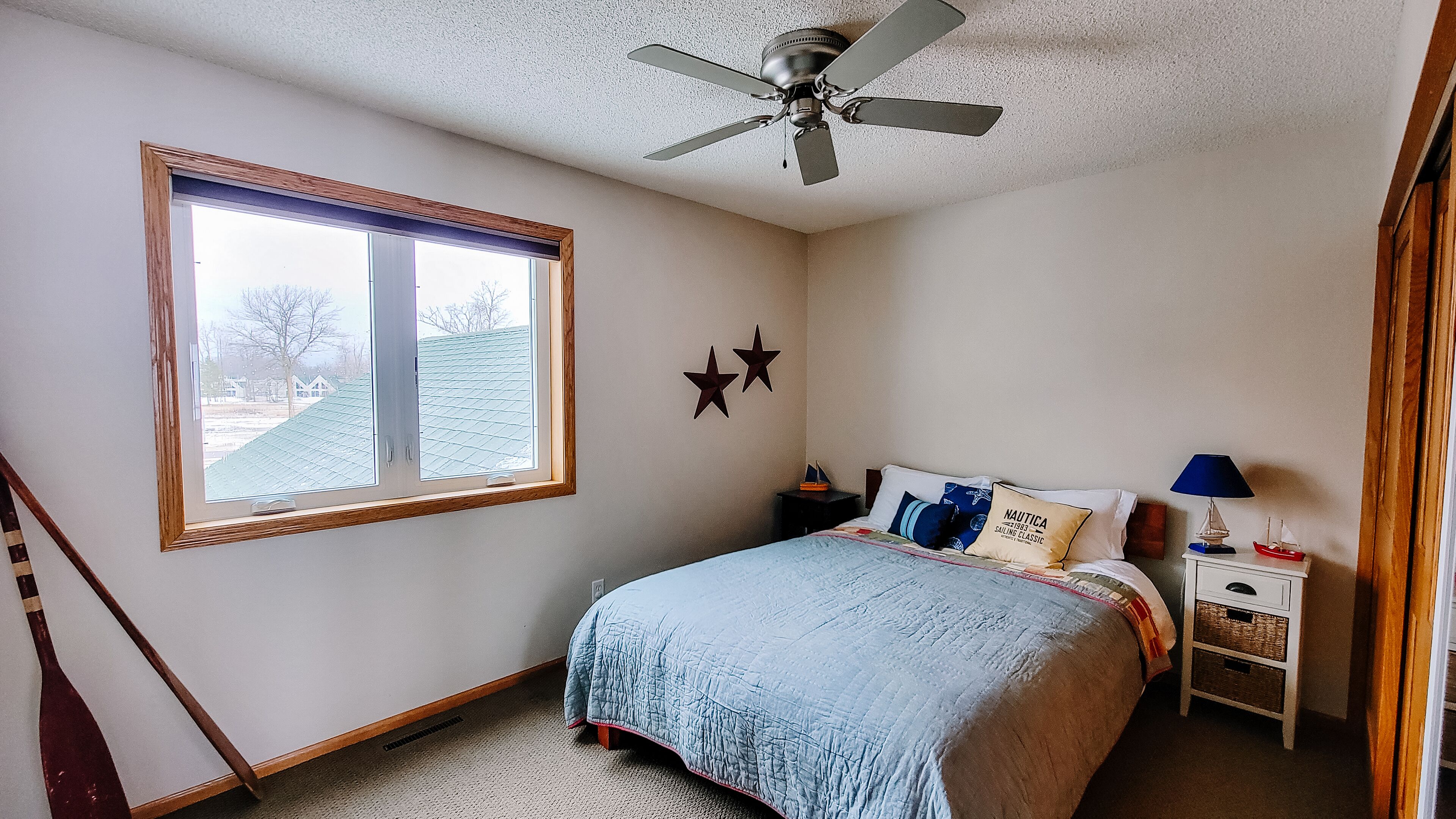 Ein Schlafzimmer mit Bett, Nachttisch, Lampe, Deckenventilator und Fenster mit Blick auf ein Haus und Bäume.