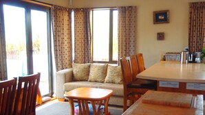 Ein Wohnzimmer mit einer Couch, Holzstühlen, einem Couchtisch, einem Esstisch und einer Küchenzeile mit Geräten.