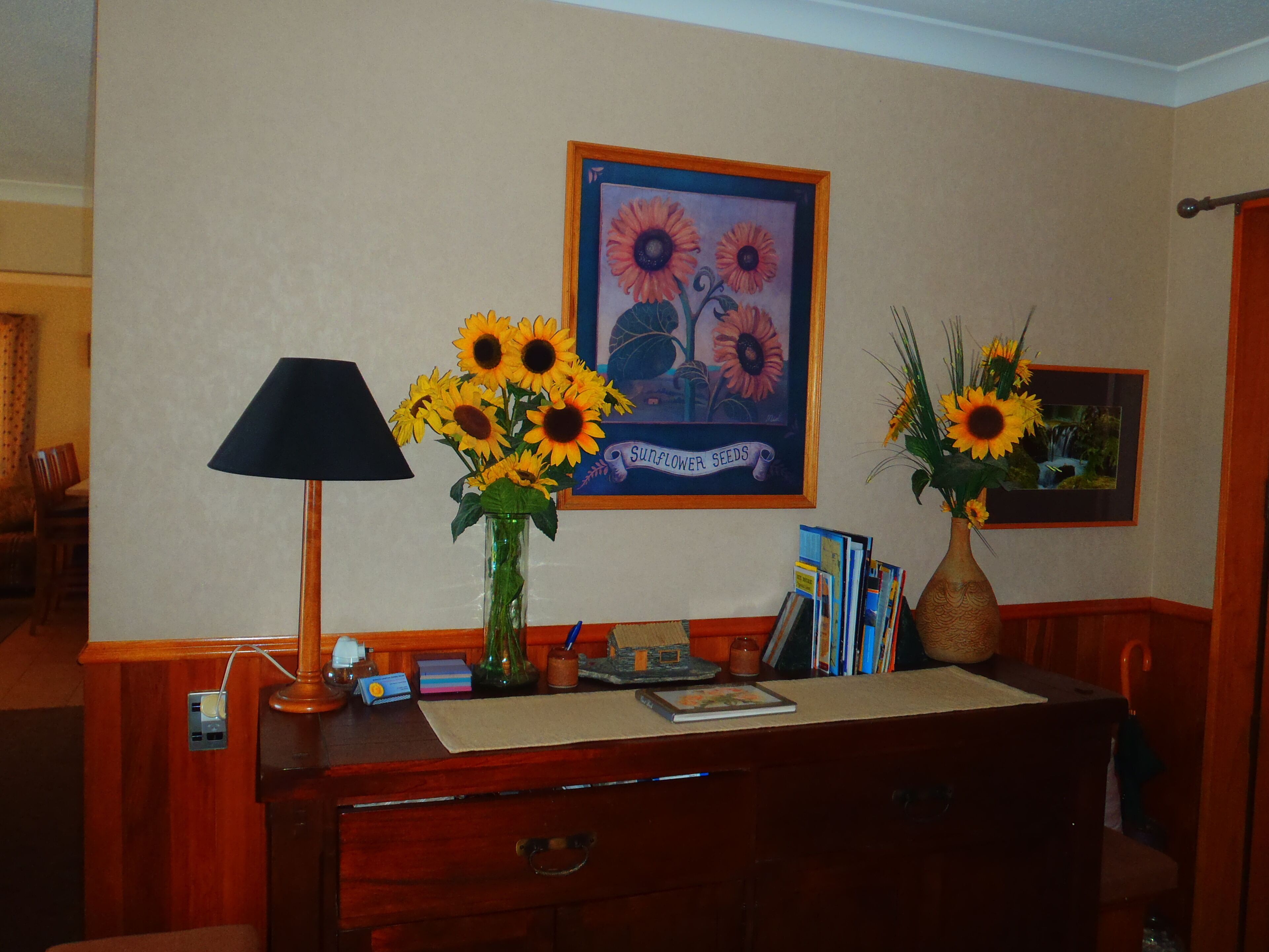 A room with a desk, a lamp, and two vases with sunflowers. There is a framed picture on the wall and a bookshelf with books and a small plant.