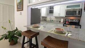 Une cuisine moderne avec des meubles blancs, un four à micro-ondes en acier inoxydable et une cuisinière. On y trouve deux tabourets en bois et une plante en pot.