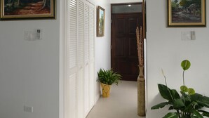 Un couloir aux murs blancs, orné de deux tableaux, avec une porte et des plantes en pot.