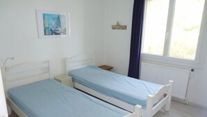 A small, clean room with two single beds, a small table, a nightstand, a window with curtains, and a wall-mounted picture.