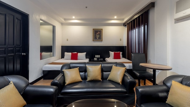 A hotel room with a black leather sofa, two beds with red and white bedding, a small table, and a large window. - Check Inn Pension Arcade (Dumaguete)