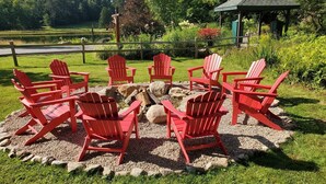 A circular seating area with red Adirondack chairs around a fire pit, set in a garden with a gazebo and a pond in the background.