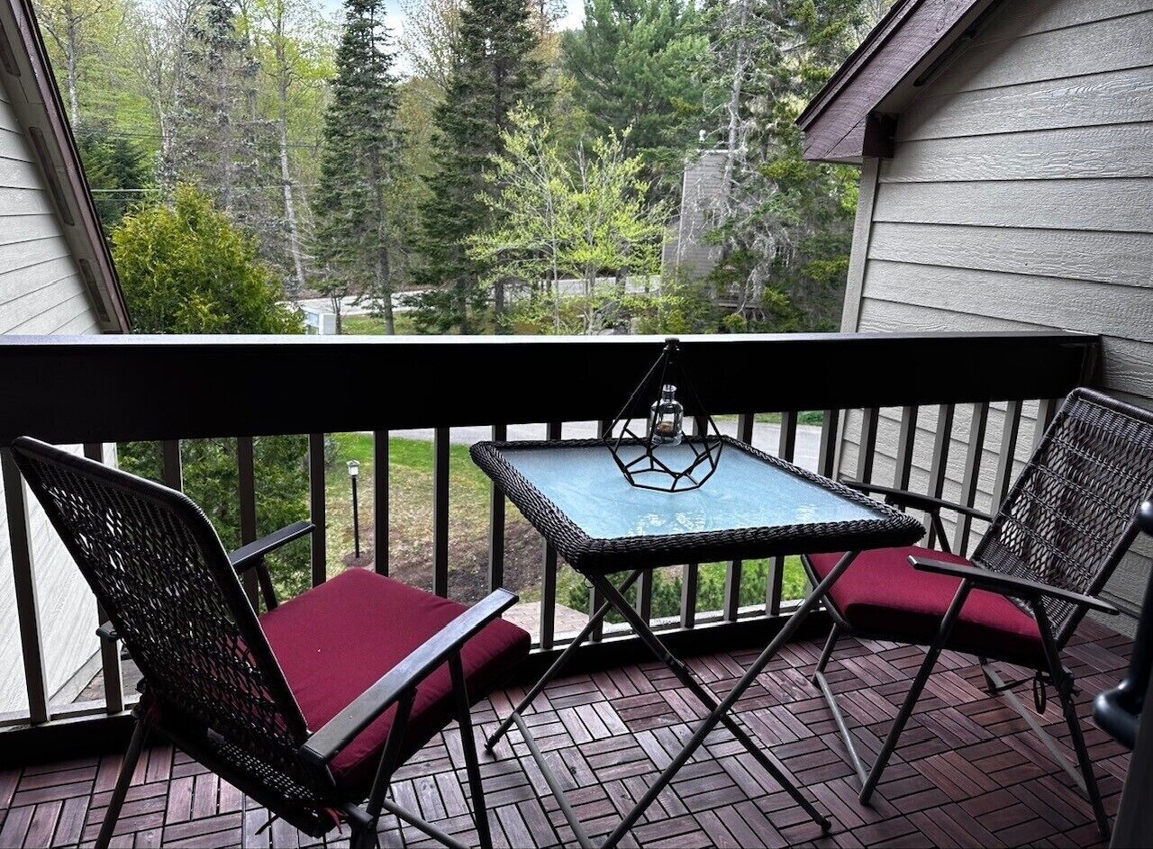 A covered outdoor area with a table and two chairs, surrounded by a wooden deck and a railing.