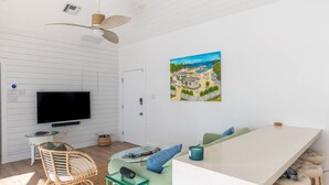 A modern living room with a flat-screen TV, wicker furniture, a green sofa, and a painting of a coastal scene. - Renovated Beach Cottage w/ Private Pool Steps Away from the Sea (North Side)