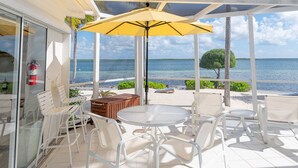A covered outdoor seating area with white chairs and a yellow umbrella, overlooking a beach with palm trees and the ocean. - Renovated Beach Cottage w/ Private Pool Steps Away from the Sea (North Side)