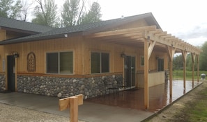 A wooden cabin with a stone foundation, a covered patio with chairs, and a wooden pergola.