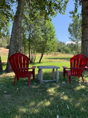 A picnic setup with two red Adirondack chairs and a wooden table in a grassy area with trees.