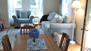 A dining area with a wooden table, chairs, and a vase of flowers. There are two sofas and a lamp in the background. - Walk to the Beach! (Middletown)