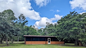 Eine einstöckige Holzhütte mit Tür und Fenstern, umgeben von einer grünen Wiese und dichten Bäumen unter einem teils bewölkten Himmel.