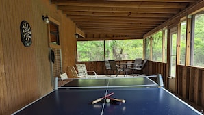 Ein Tischtennisplatte mit Netz, eine Dartscheibe und ein Billardtisch in einem überdachten Bereich mit Holzwänden und großen Fenstern.