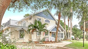 A two-story house with a balcony, surrounded by tropical plants and a well-maintained lawn. - Tropical Chalet w/ 3 King Bedrooms. 3 patios, and an Above-Ground Pool! (Sebastian)